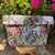 Floral-patterned fabric basket with tassels on a wooden stand, surrounded by pink flowers.