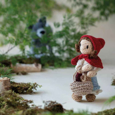Crocheted doll in a red hood and white dress with a basket, standing in a forest setting | Yarn Worx