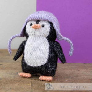 Knitted penguin toy with a purple hat on a wooden surface with snow-like material | Yarn Worx
