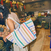 Person holding a striped tote bag in a market setting with produce and 'Patateria Macia' sign | Yarn Worx