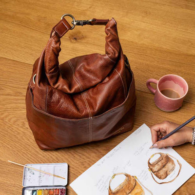 Brown leather handbag on a wooden table with a person drawing and a pink mug | Yarn Worx