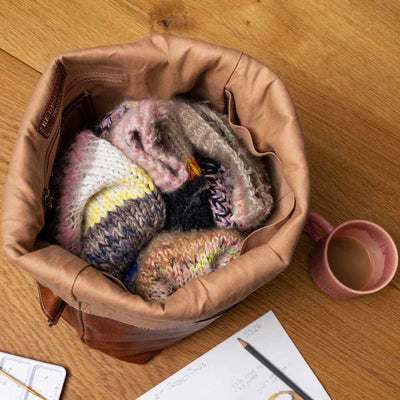 Brown leather bag filled with knitted items on a wooden surface with a pink mug and notebook | Yarn Worx
