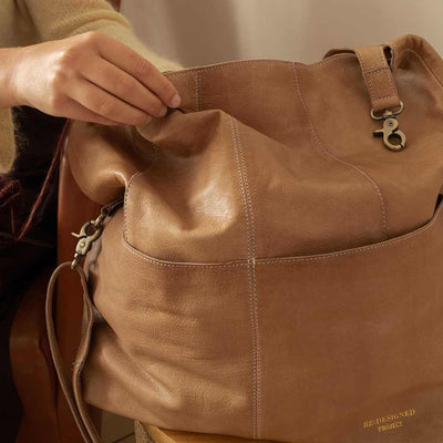 Brown leather bag with a person's hand opening it, on a neutral background | Yarn Worx
