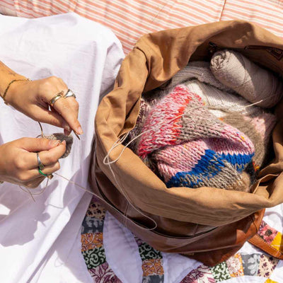 Person knitting with yarn and needles, surrounded by a basket of yarn and knitting materials on a patterned surface | Yarn Worx