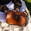 A brown leather doctors-style handbag with silver hardware, held by a person sitting outdoors on a quilted blanket. The person is wearing a dark brown knit sweater and white pants, with green grass visible in the background | Yarn Worx