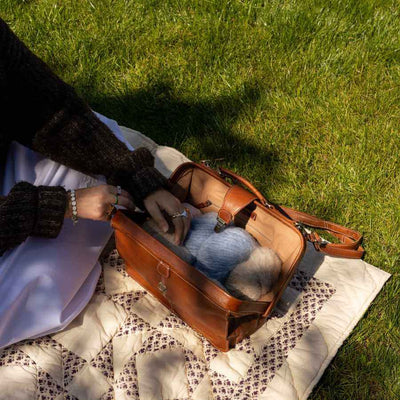 A brown leather shoulder bag is placed on a quilted blanket on grass, with a person's hands reaching into the bag to retrieve a blue knit item. The scene is shot in natural outdoor sunlight | Yarn Worx