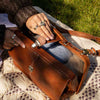 A brown leather shoulder bag with silver-tone hardware, shown being held open by two hands wearing rings, resting on a patterned blanket outdoors on grass | Yarn Worx