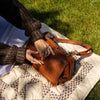 A person outdoors on a quilted blanket over grass, holding a brown leather shoulder bag. The person wears a dark brown knit sweater and rings, and the bag has a long strap and metal hardware | Yarn Worx