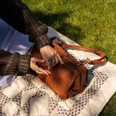 A person outdoors on a quilted blanket over grass, holding a brown leather shoulder bag. The person wears a dark brown knit sweater and rings, and the bag has a long strap and metal hardware | Yarn Worx