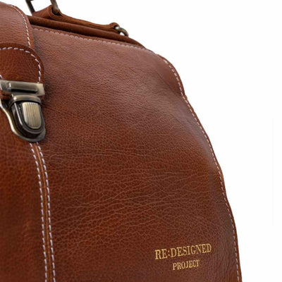 Close-up of a brown leather bag with white stitching, a metal clasp, and gold embossed text reading 'RE-DESIGNED PROJECT' on a white background | Yarn Worx