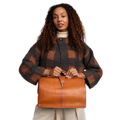 A woman with long curly brown hair holds a tan leather doctors-style bag in front of her torso. She is wearing a dark brown and brown plaid fuzzy cardigan over a cream turtleneck, and the background is a clean white studio backdrop | Yarn Worx