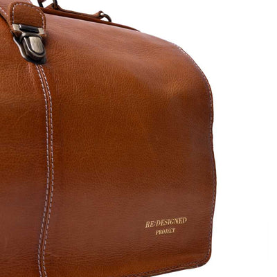 Close-up of a brown leather bag corner with white contrast stitching, a metal clasp, and gold embossed text reading 'RE-DESIGNED PROJECT' on a white background | Yarn Worx