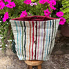 Striped fabric planter with a button on top, placed outdoors with pink flowers in the background.