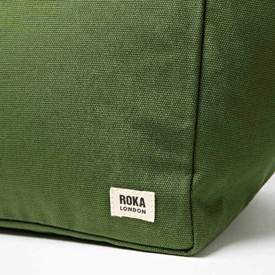 Close-up of an olive green canvas tote bag corner with a white rectangular ROKA LONDON brand label sewn onto the side | Yarn Worx
