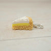 Keychain shaped like a slice of cake with white frosting and yellow filling on a light wooden surface.