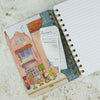 A spiral-bound notebook with a colorful illustrated cover featuring a street scene, shown open with lined pages and a receipt tucked into the pocket, placed on a light textured surface | Yarn Worx