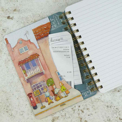 A spiral-bound notebook with a colorful illustrated cover featuring a street scene, shown open with lined pages and a receipt tucked into the pocket, placed on a light textured surface | Yarn Worx