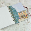 An open spiral-bound notebook lying on a textured light surface. The left page is lined and blank, while the right cover shows a teal pattern of small illustrated buildings and a colourful street-art style storefront illustration with a yellow car | Yarn Worx