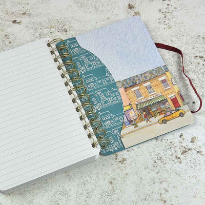 An open spiral-bound notebook lying on a textured light surface. The left page is lined and blank, while the right cover shows a teal pattern of small illustrated buildings and a colourful street-art style storefront illustration with a yellow car | Yarn Worx